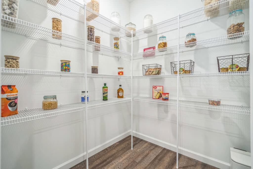 Spacious pantry with white wire shelves holding jars, baskets, and boxes of food items, against white walls and wood flooring.