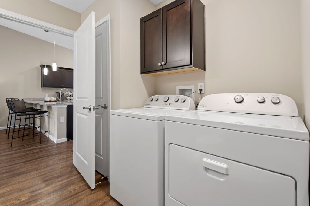 Laundry area with a white washing machine and dryer, brown cabinets above, hardwood floors, and a view of the kitchen with bar stools in the background.