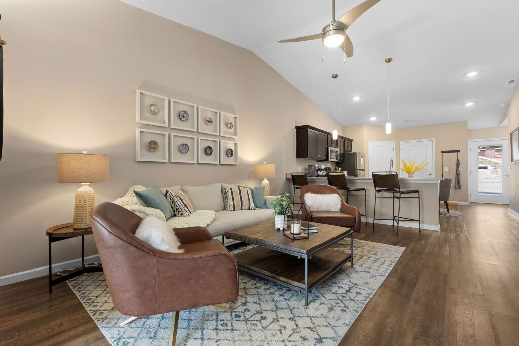 Open-concept living room with brown leather chairs, beige sofa, coffee table, rug, and kitchen with barstools in the background under pendant lights.