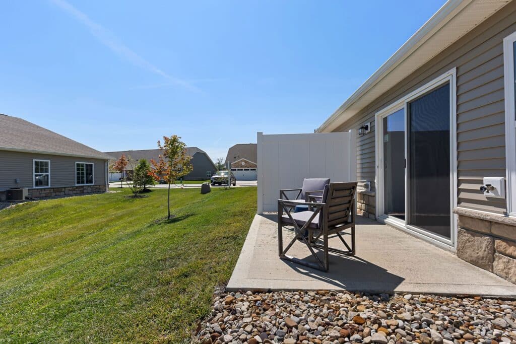 A small concrete patio with two chairs and a white privacy screen sits beside a house, overlooking a grassy backyard and neighboring homes on a clear day.