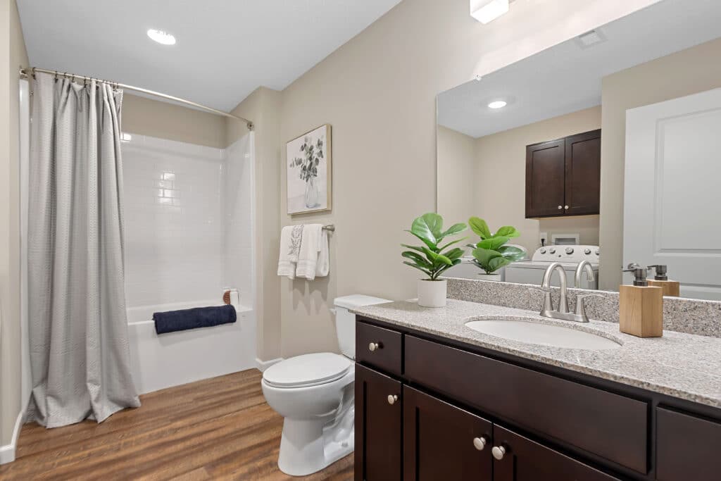 Modern bathroom with a shower-tub combo, white curtain, granite countertop, dark wood vanity, potted plant, towels, and a partial view of a laundry area in the background.