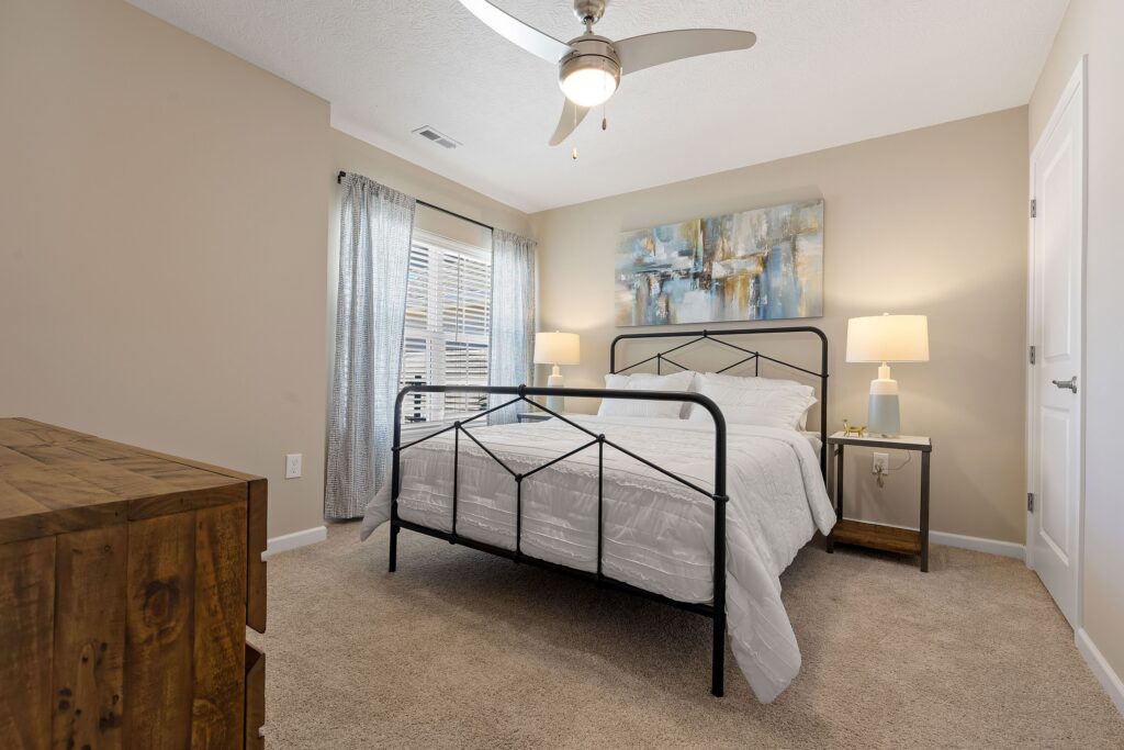 A neatly made bed with white bedding, black metal frame, two nightstands with lamps, abstract artwork above the bed, and a wooden dresser in a beige carpeted bedroom.
