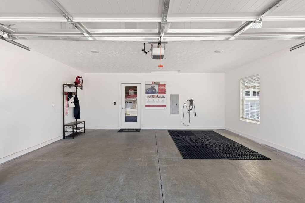Clean, spacious garage with concrete floor, wall-mounted shelving, a door, a window, posters, and rubber floor mats. Ceiling-mounted garage door opener is visible.