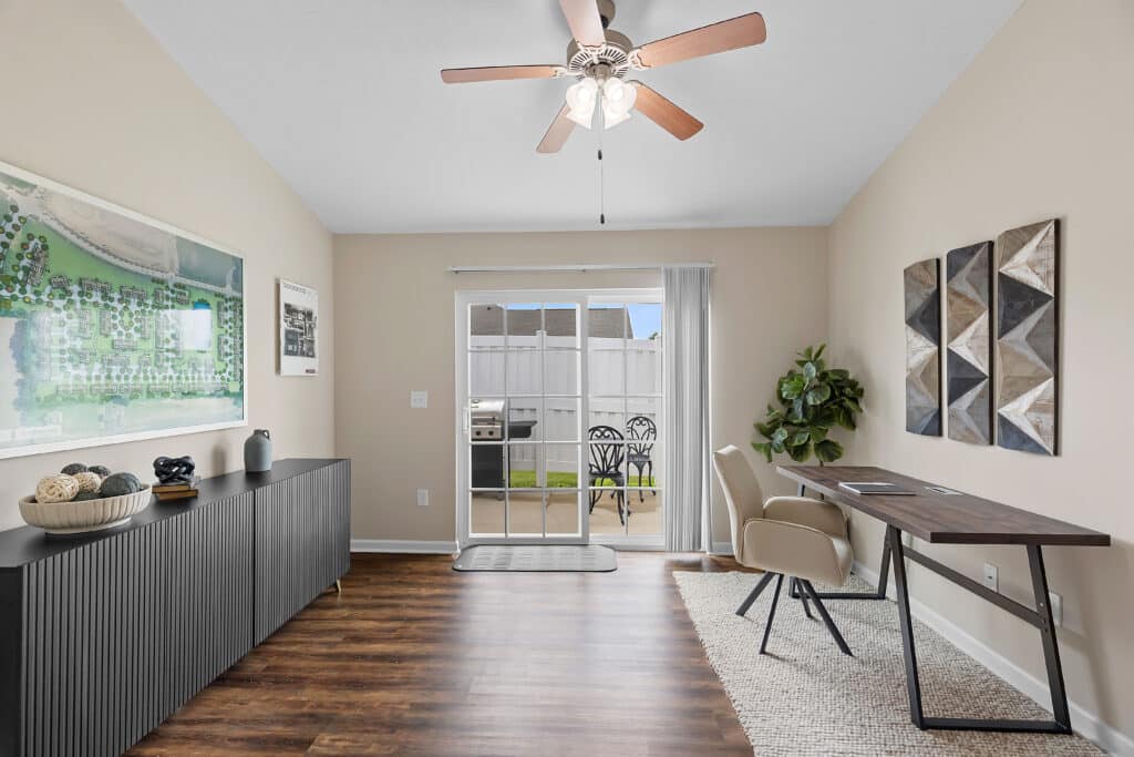 Modern home office with a desk, chair, and decor; sliding glass doors open to a patio with outdoor seating and a grill. Neutral walls and wood flooring are visible.