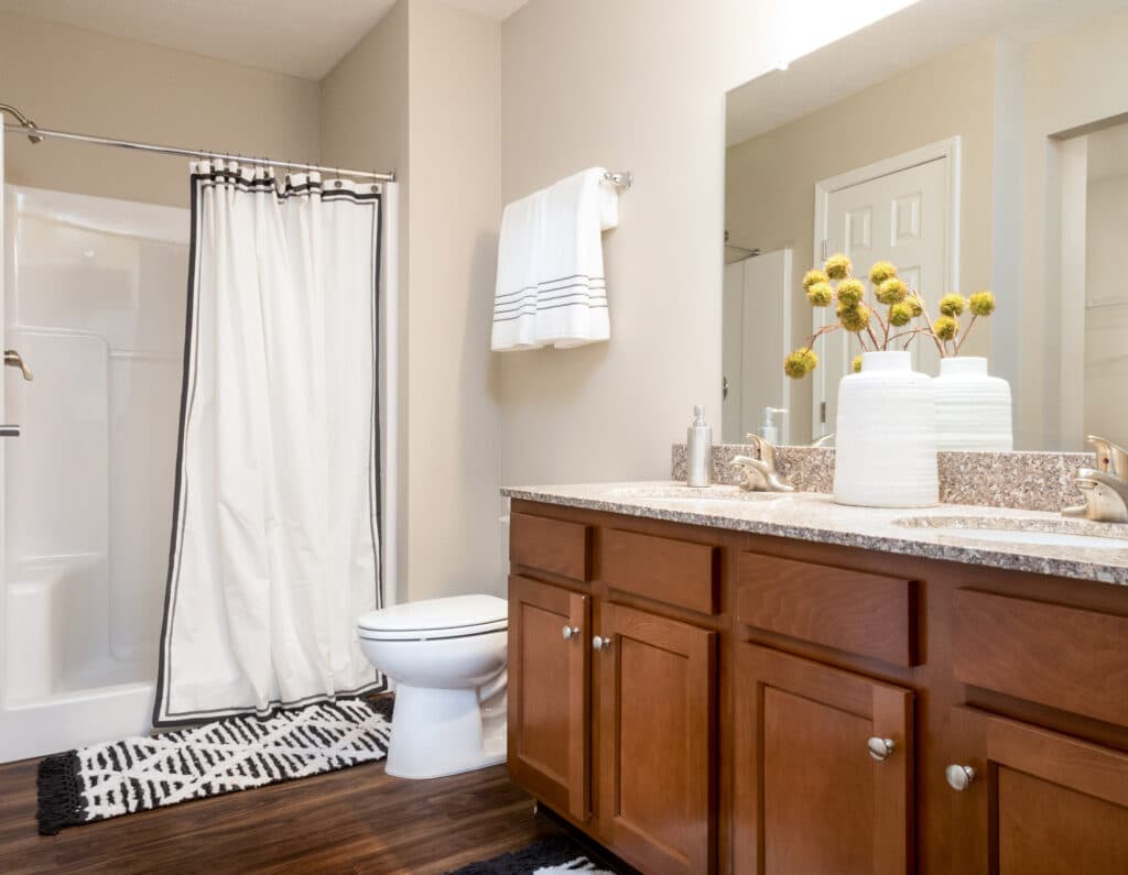 A bathroom with a shower and white curtain, a toilet, double sink vanity with granite countertop, wooden cabinets, and decorative vases with yellow flowers.