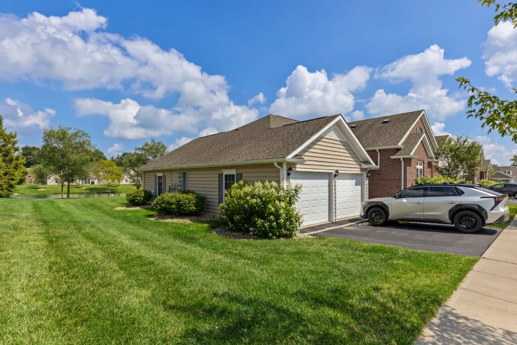 A single-story house with a two-car garage, a white SUV parked in the driveway, and a well-kept green lawn under a partly cloudy sky.