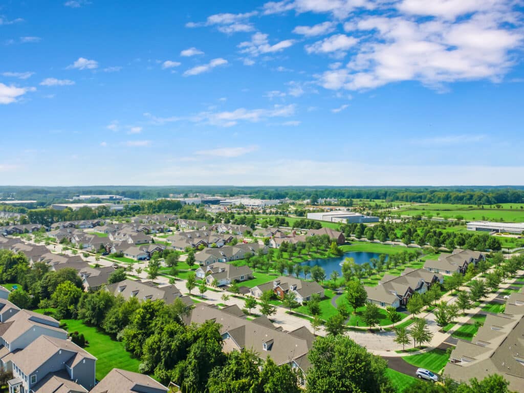 Aerial view of a suburban neighborhood with rows of houses, green lawns, a pond, and surrounding fields under a partly cloudy sky.