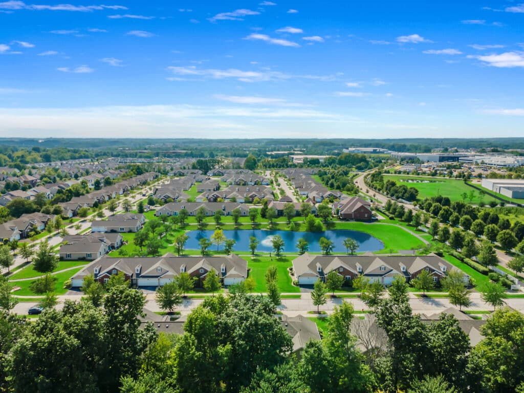 Aerial view of a suburban neighborhood with rows of houses, green lawns, trees, and several ponds under a clear blue sky.