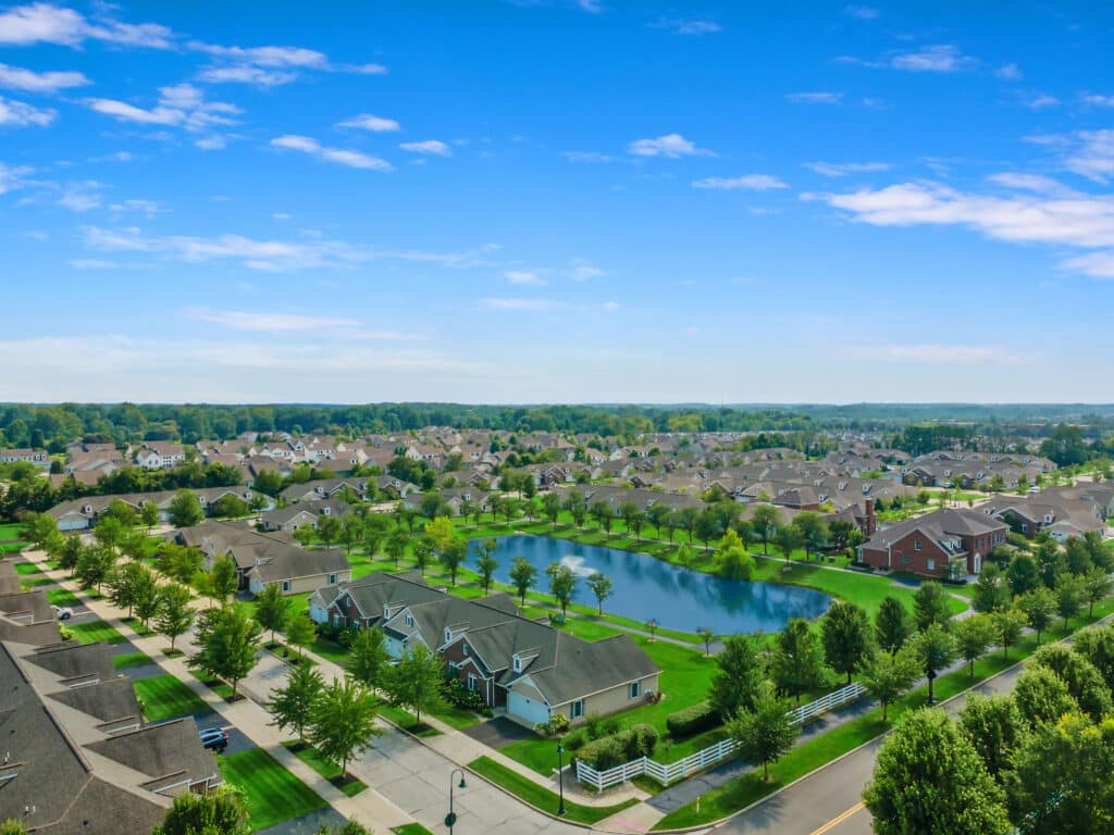 Aerial view of a suburban neighborhood with rows of houses, tree-lined streets, a pond, and a clear blue sky.