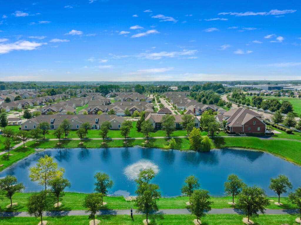 A suburban neighborhood with houses, a pond with a fountain, and green trees under a blue sky with scattered clouds.