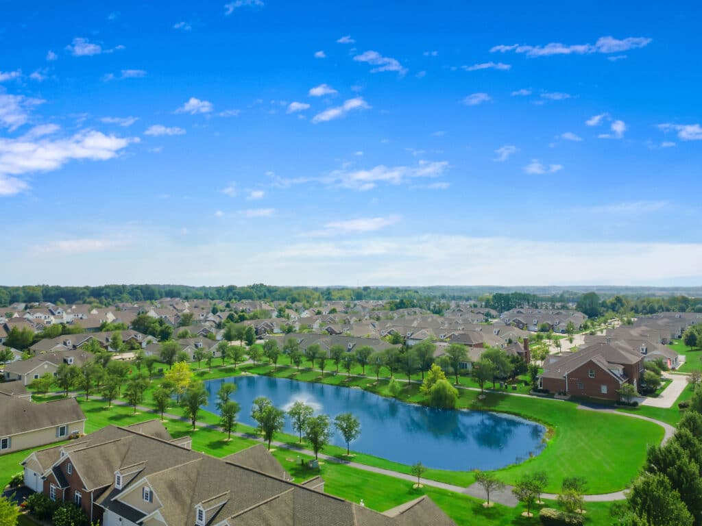 Aerial view of a suburban neighborhood with rows of houses, a small pond, and green lawns under a blue sky with scattered clouds.