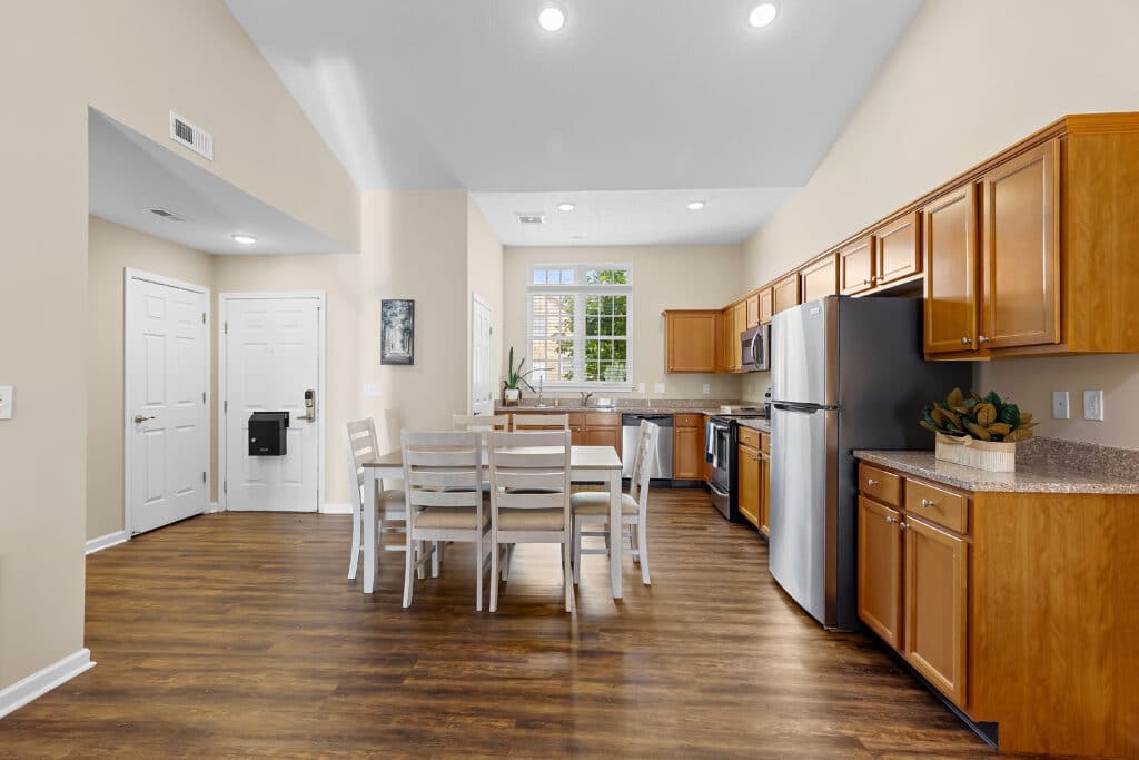 Open kitchen and dining area with wooden floors, light wood cabinets, stainless steel appliances, and a white dining table with six chairs. White front door and windows in the background.