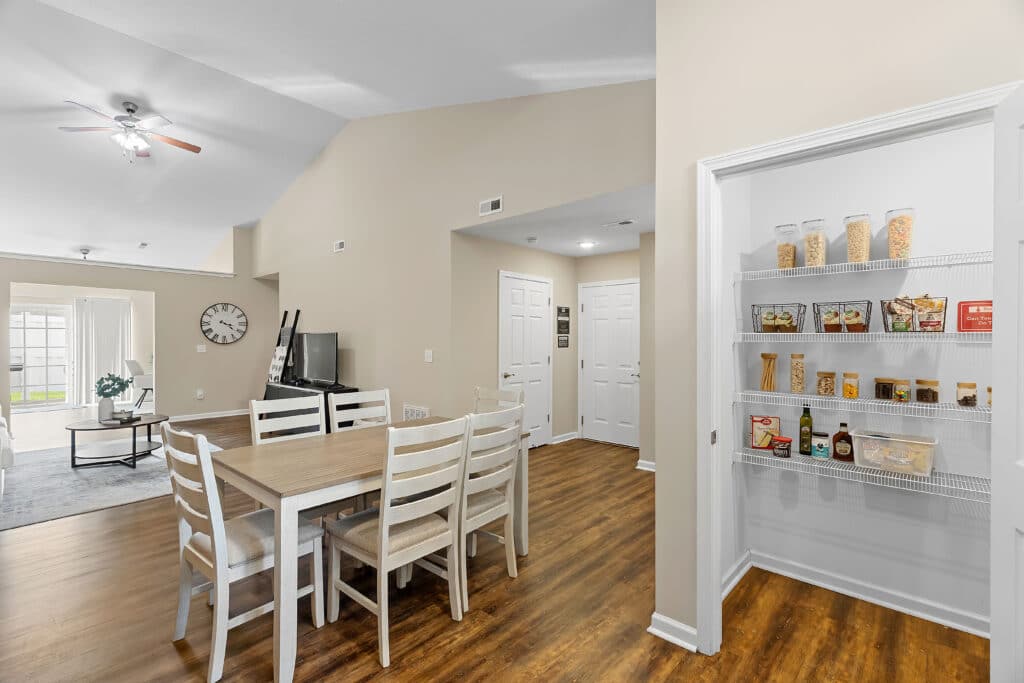 A dining area with a wooden table and six chairs, an adjacent open pantry with shelves of food items, and a view into a living room with a TV and clock on the wall.