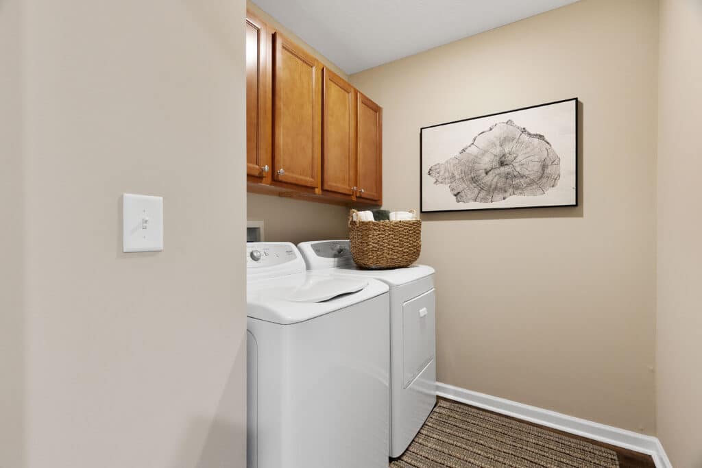 Laundry room with a white washer and dryer, wooden cabinets above, a wicker basket on top, beige walls, framed tree ring artwork, and a striped floor mat.
