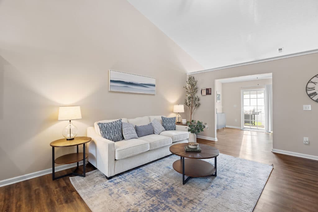 A living room with a beige sofa, blue and gray cushions, two round wooden tables with lamps, a wall art piece, potted plant, and a rug on hardwood flooring.