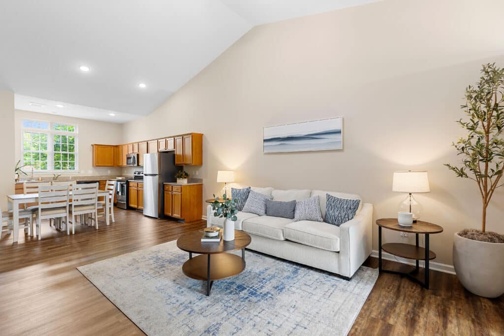Open-concept living room and kitchen with wood cabinetry, stainless steel appliances, a dining table, beige sofa, area rug, and potted plant; neutral colors and natural light.