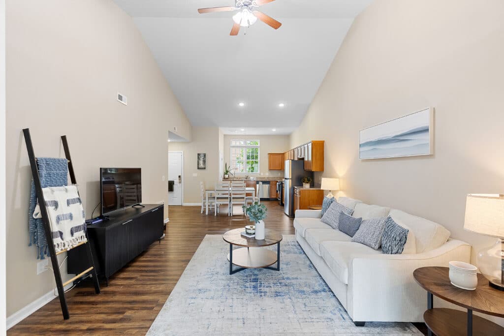 Open-concept living room with a cream sofa, blue accents, TV stand, and kitchen with wooden cabinets visible in the background, featuring high ceilings and wood flooring.