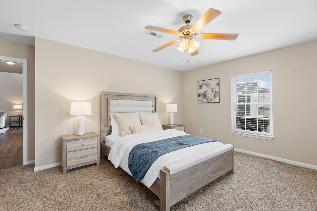 A bedroom with a wooden bed, two nightstands with lamps, beige walls, carpeted floor, a ceiling fan, and a window with blinds.