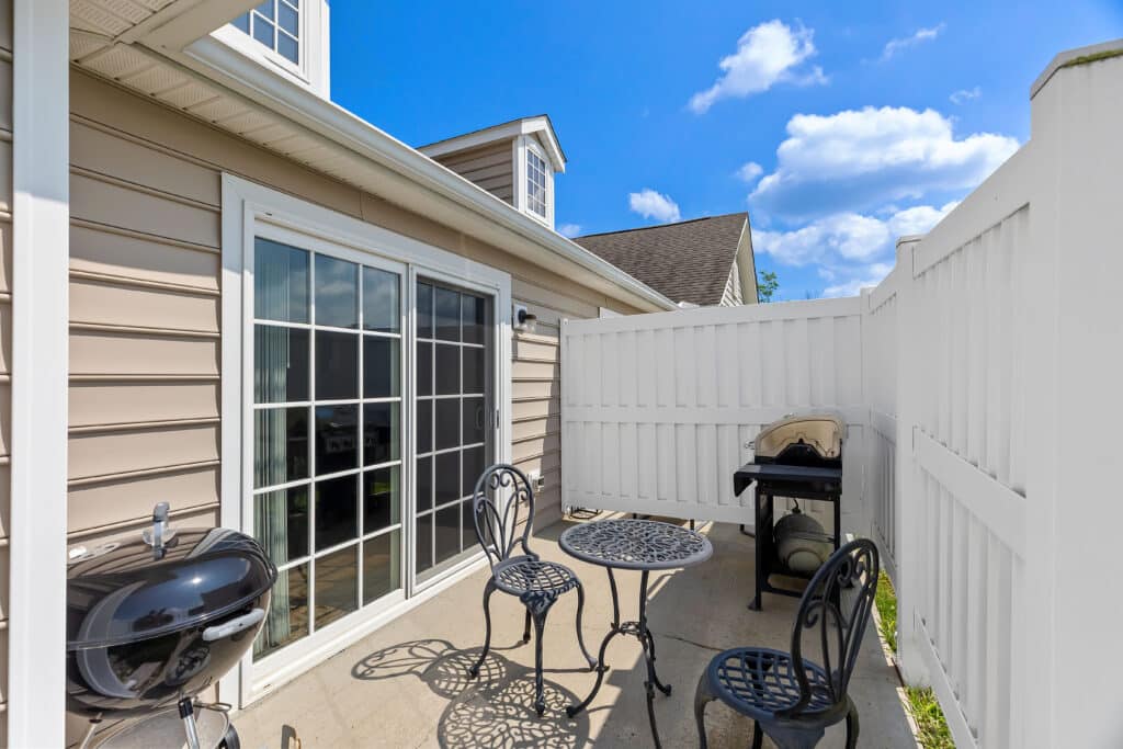 Small patio area with a round black metal table, three chairs, a charcoal grill, a gas grill, beige siding, sliding glass doors, and a white privacy fence under a blue sky.