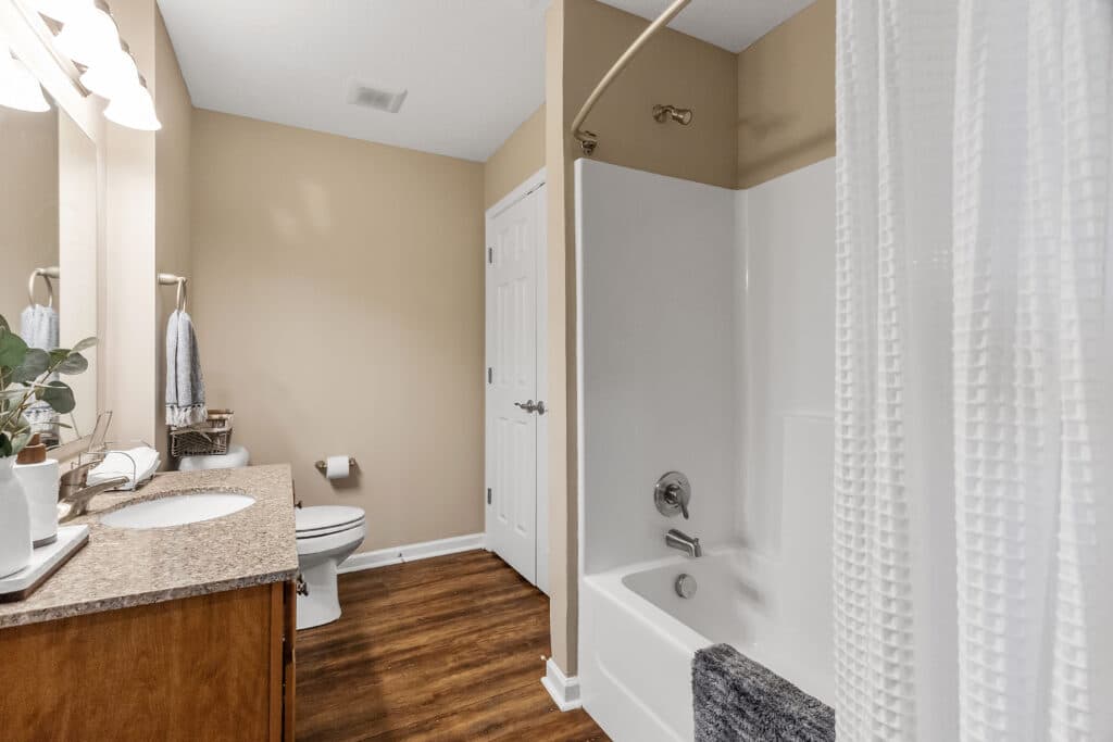 A bathroom with a white bathtub and shower, a toilet, a single-sink vanity with a granite countertop, and light brown walls and floor.