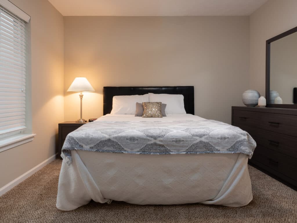 A neatly made bed with a patterned blanket, decorative pillows, a lamp on a nightstand, and a dresser with a mirror and vases in a carpeted bedroom.
