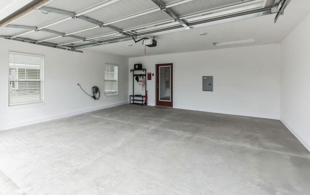 Empty two-car garage with white walls, gray concrete floor, two windows, a red door, and ceiling-mounted garage door opener. Minimal shelving and equipment on the wall.