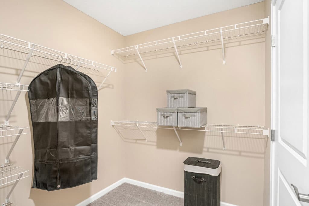 Empty walk-in closet with beige walls, white wire shelves, a black garment bag hanging, two gray storage boxes, and a black laundry hamper on carpeted floor.