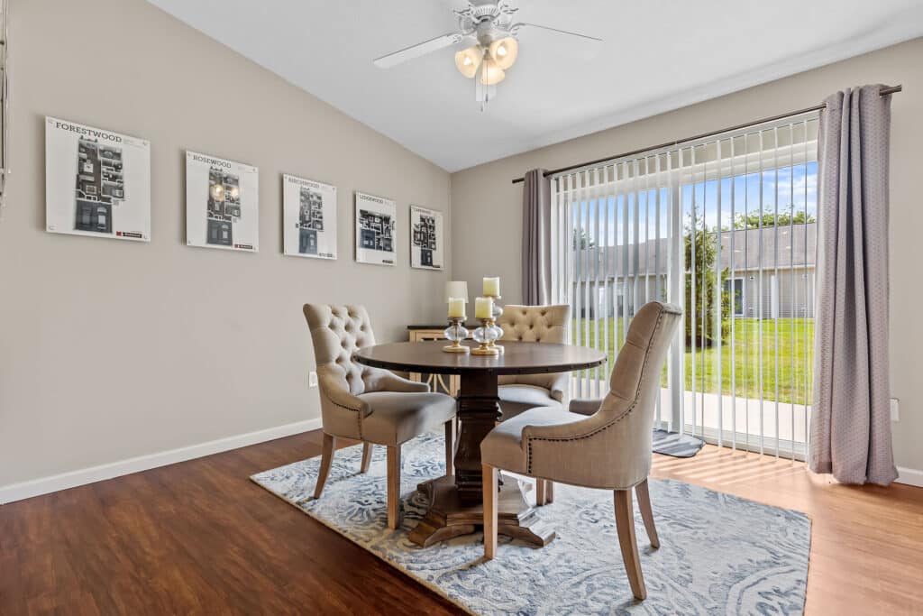 A dining area with a round table, four upholstered chairs, a rug, wall art, and sliding glass doors with vertical blinds overlooking a grassy yard.