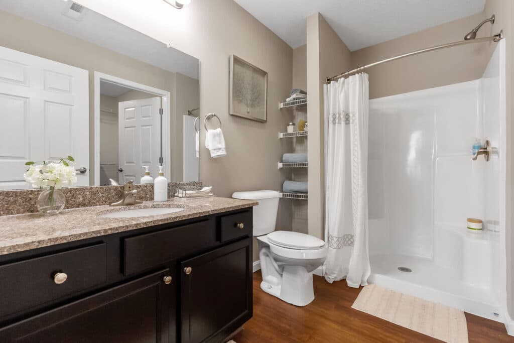 Modern bathroom with dark wood vanity, granite countertop, toilet, walk-in shower with curtain, shelving unit with towels, and a mirror above the sink.