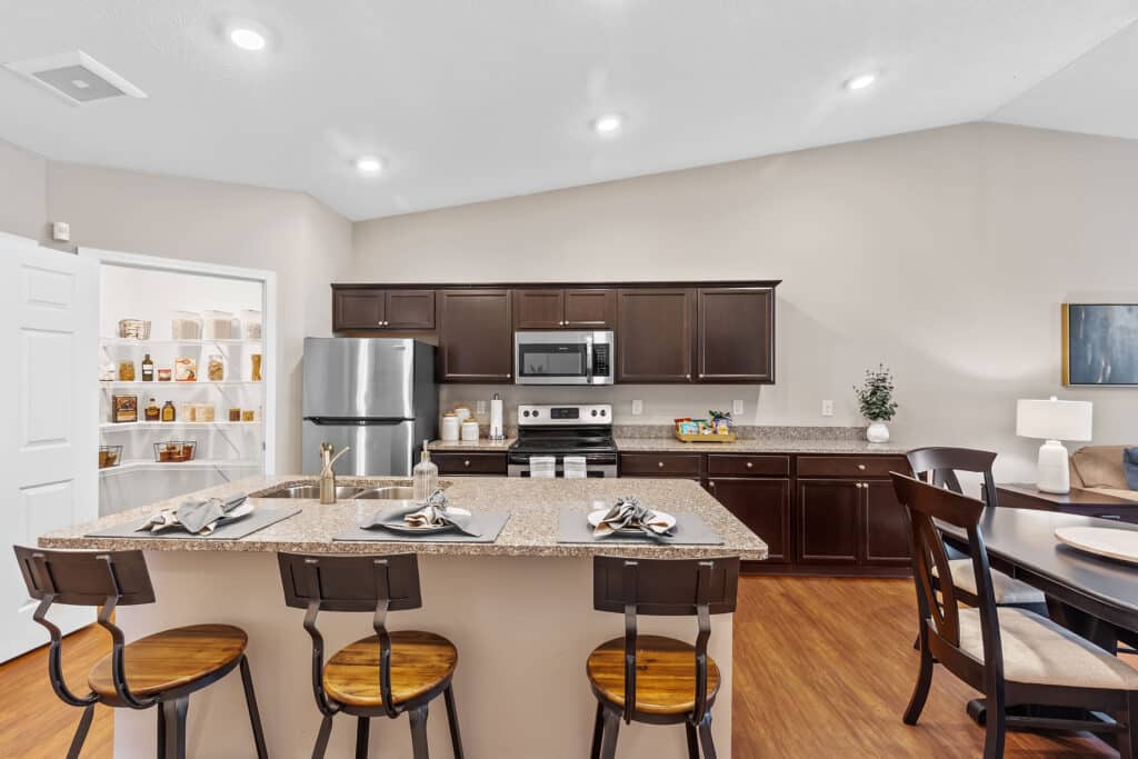 Modern kitchen with dark wood cabinets, stainless steel appliances, an island with seating for three, and an open pantry door revealing organized shelves.