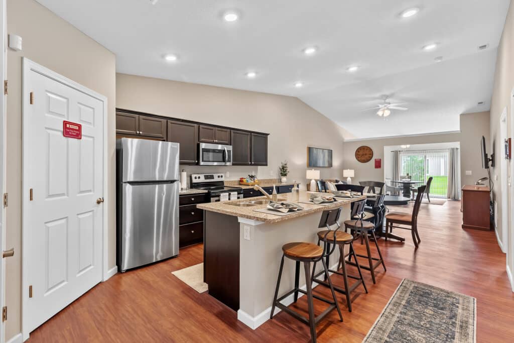 Modern open-concept kitchen and dining area with stainless steel appliances, dark cabinets, a breakfast bar with four stools, and a dining table; sliding doors reveal a yard.