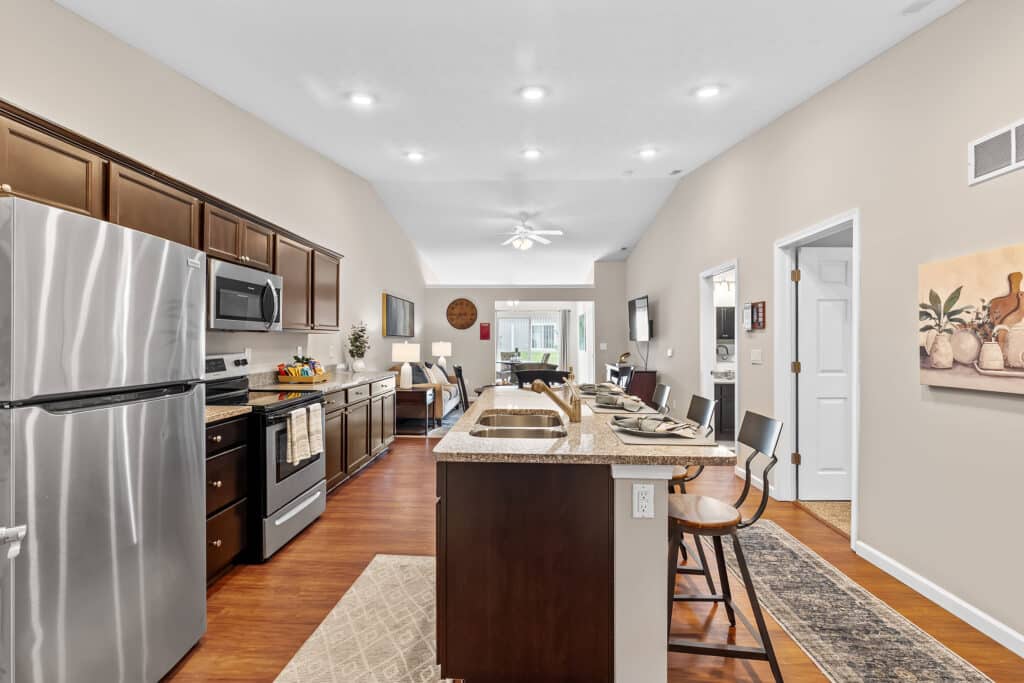 Modern kitchen with stainless steel appliances, dark wood cabinets, a central island with bar stools, and an open view into a bright living area.