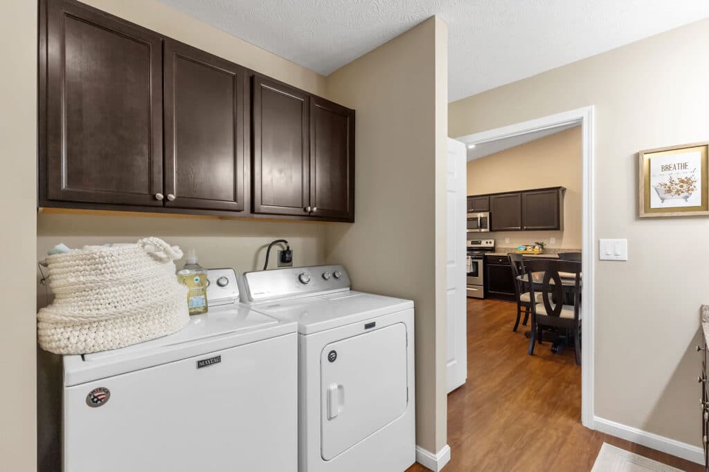 Laundry room with a white washer and dryer, dark wood cabinets above, and a woven basket on top; view into a kitchen and dining area with wood flooring.