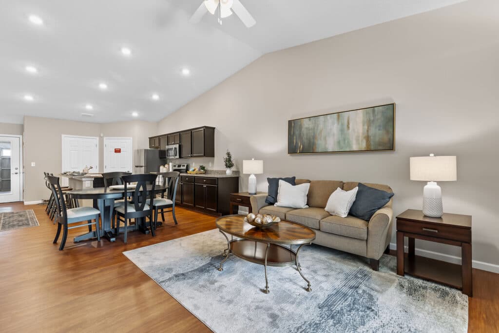Open-concept living room and kitchen with neutral decor, a brown sofa, coffee table, dining set, dark cabinets, and wood flooring under recessed lighting.