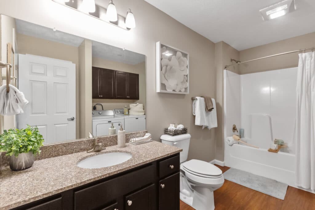 Modern bathroom with a granite countertop, dark wood cabinets, a toilet, tub with shower curtain, and a laundry area with washer and dryer in the background.