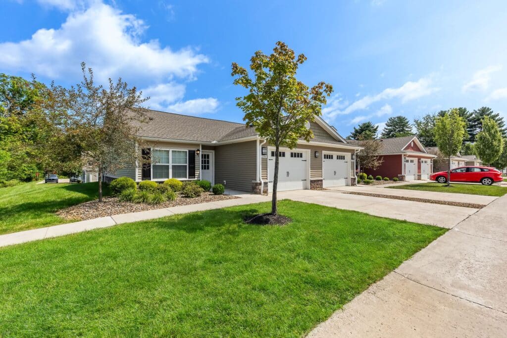 Single-story duplex homes with attached garages, well-maintained lawns, and a red car parked in a driveway under a blue sky with scattered clouds.