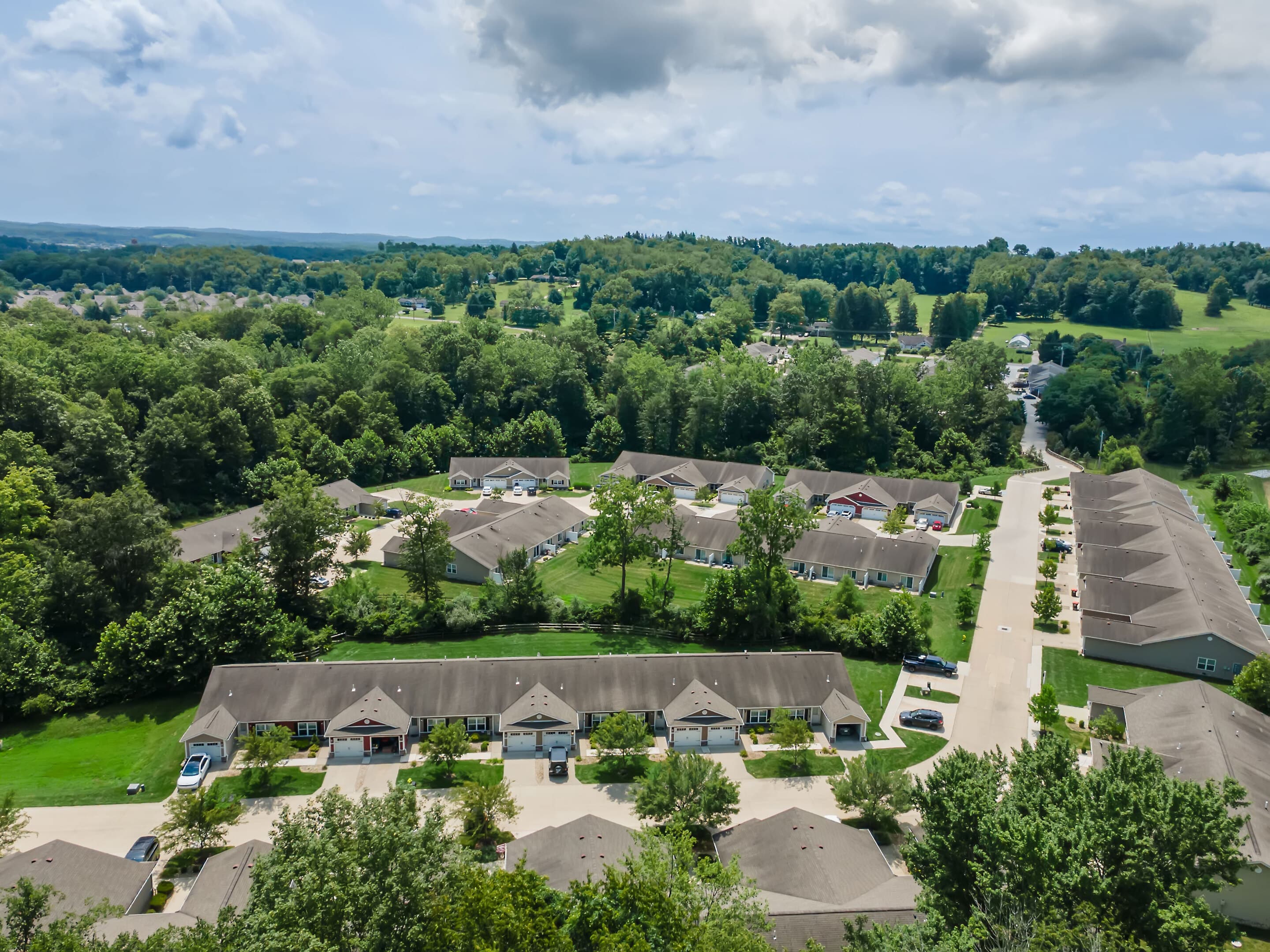 Aerial view of a suburban neighborhood with single-story houses, driveways, green lawns, trees, and a forested area in the background under a cloudy sky.
