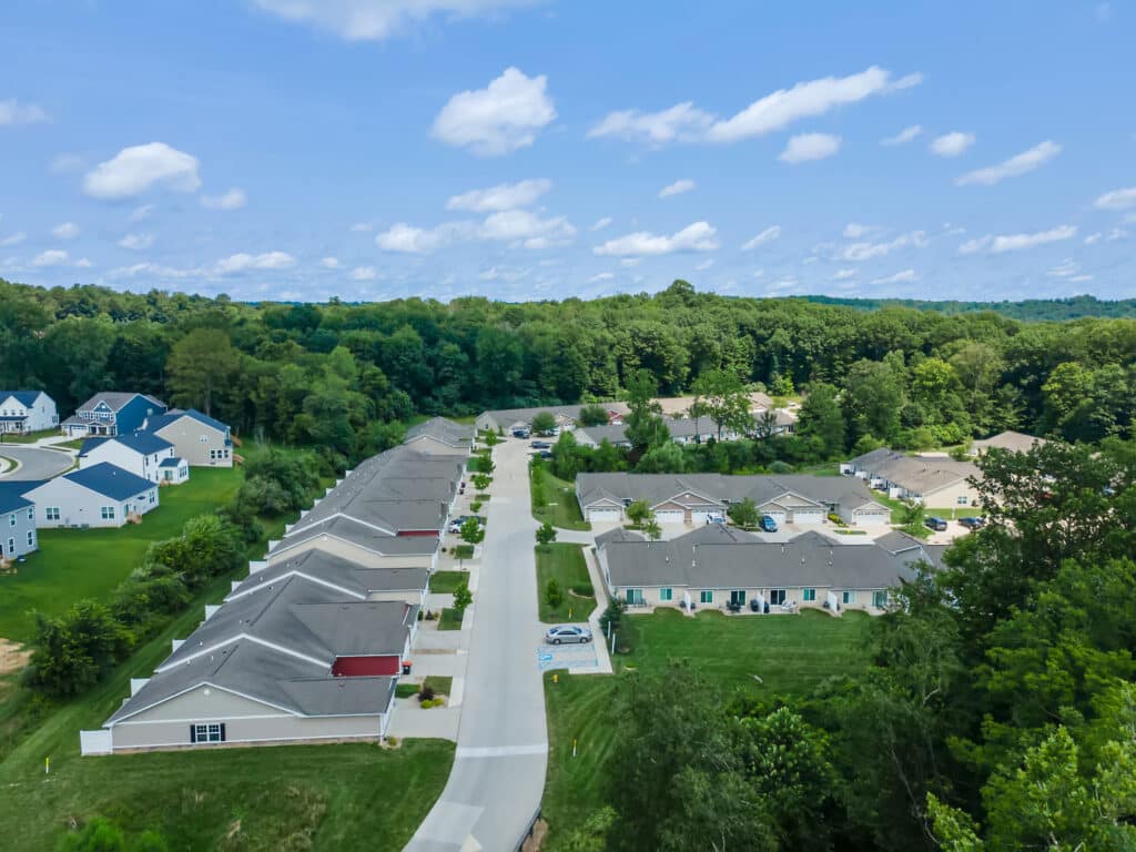 Aerial view of a suburban neighborhood with rows of single-story houses, green lawns, trees, and a street running through the center.