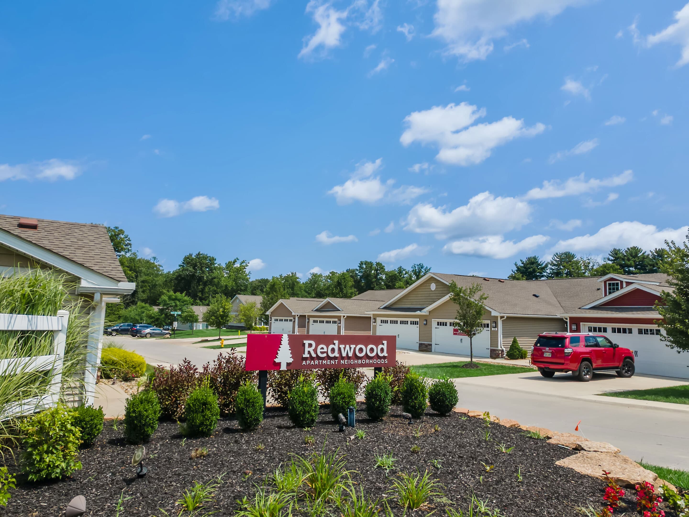 A red sign reading "Redwood Apartment Neighborhood" stands in front of landscaped bushes, with single-story houses and a red SUV in the background under a blue sky.