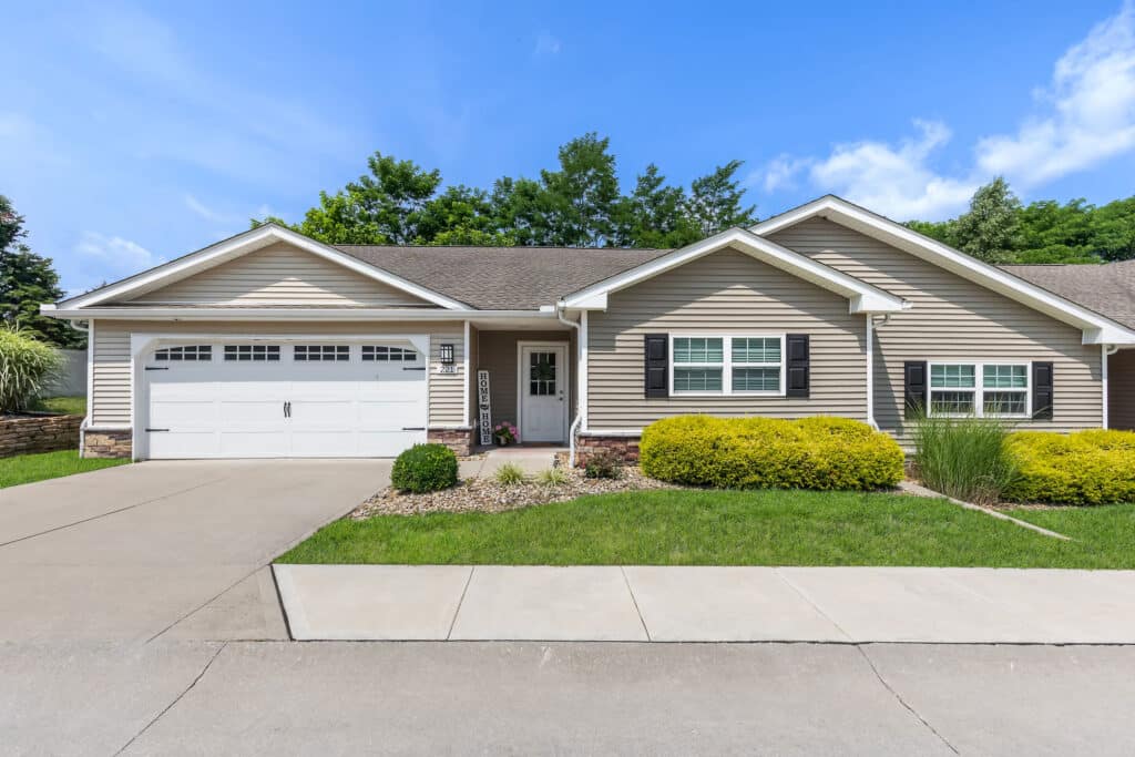 Single-story suburban house with beige siding, white trim, a two-car garage, manicured lawn, shrubs, and a concrete driveway under a clear blue sky.