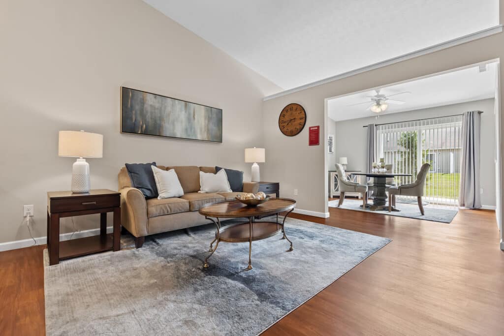 A living room with a couch, two side tables with lamps, a coffee table, wall art, and a clock, adjoining a dining area with a table, chairs, and glass doors to a patio.