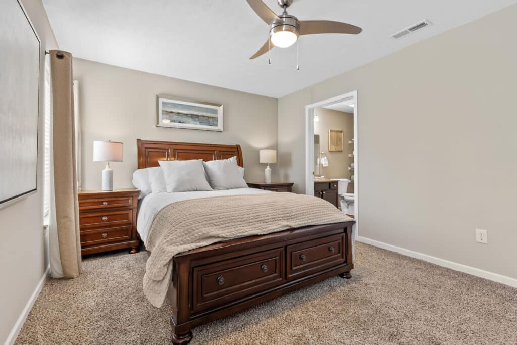A neatly made bed with a wooden frame is centered in a carpeted bedroom, with two nightstands, lamps, a ceiling fan, and an open door leading to a bathroom.