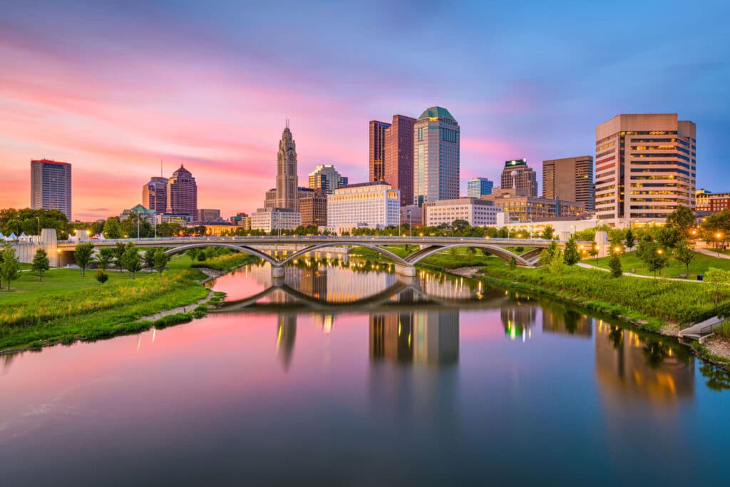 Downtown city skyline with modern buildings at sunset, a bridge crossing a river, and green spaces along the waterfront.