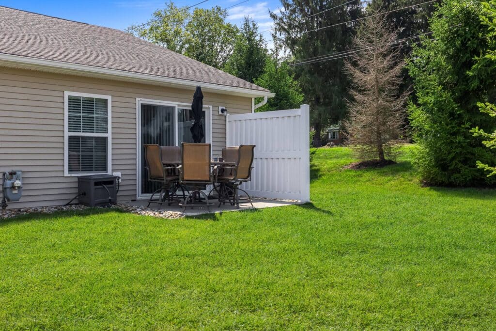 A small concrete patio with a table, six chairs, and a closed umbrella is next to a sliding glass door on the back of a beige house, surrounded by green grass and trees.