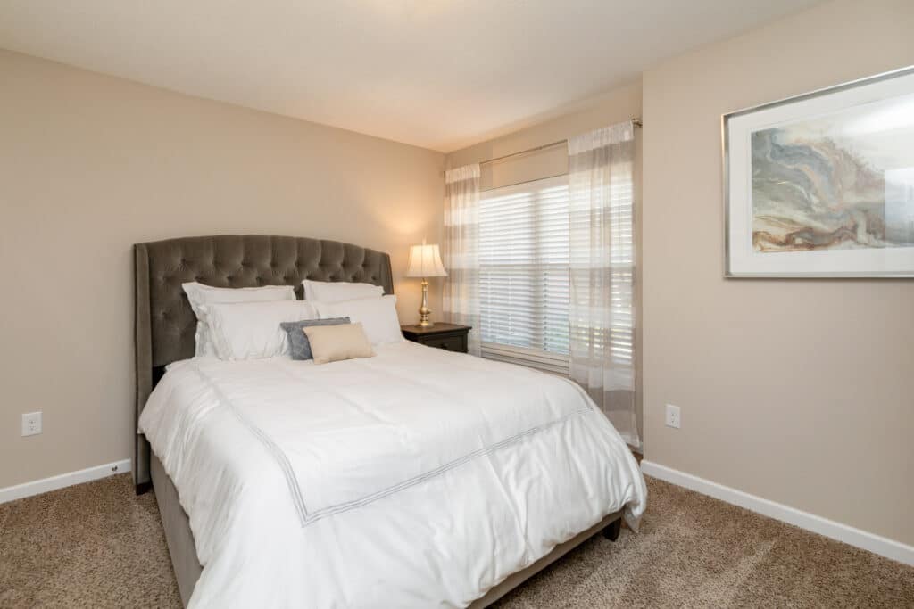 A neatly made bed with white bedding in a carpeted bedroom, featuring a tufted headboard, nightstand with a lamp, window with sheer curtains, and a framed abstract artwork.