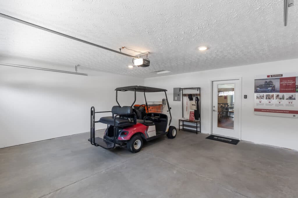 A red and black golf cart parked inside a clean, spacious garage with white walls and a concrete floor. A door and informational posters are visible on the right wall.