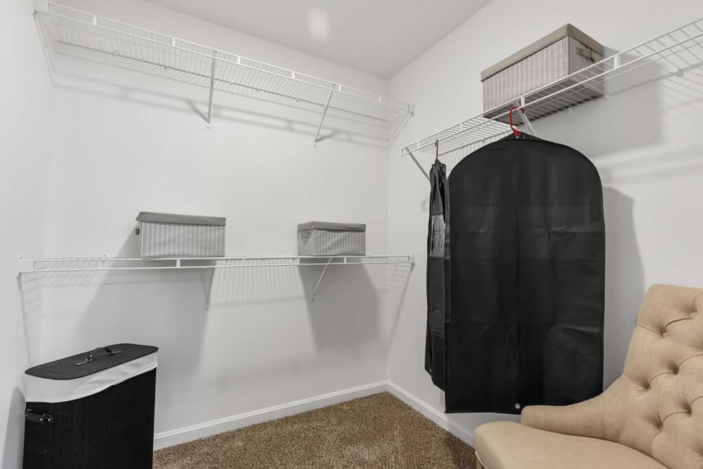 A walk-in closet with wire shelving, a laundry hamper, storage bins, a hanging garment bag, and a beige upholstered chair on carpeted flooring.