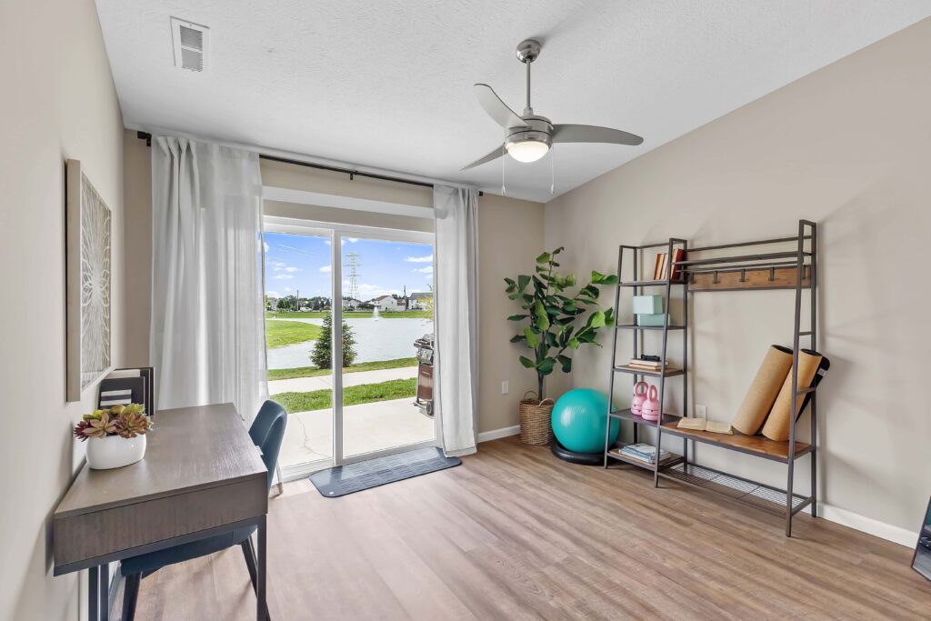 Bright room with a desk, chair, exercise equipment, shelving, and a plant, featuring wood flooring and sliding glass doors that open to a view of a lake.