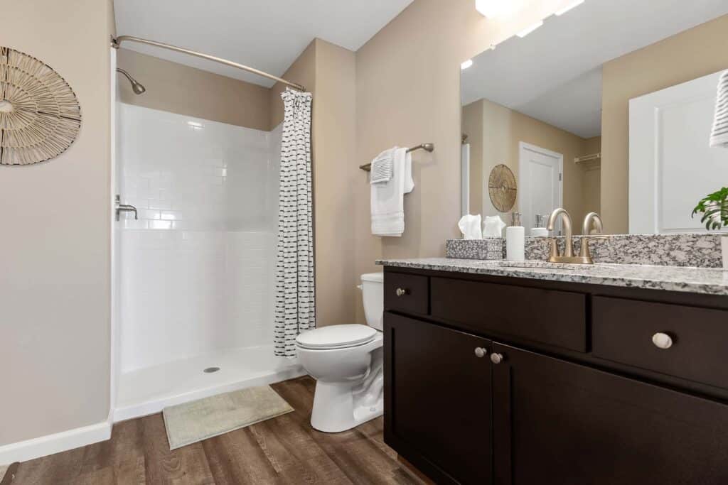 Modern bathroom with a walk-in shower, patterned curtain, toilet, dark vanity with granite countertop, and wood-style flooring. Towels and toiletries are neatly arranged.