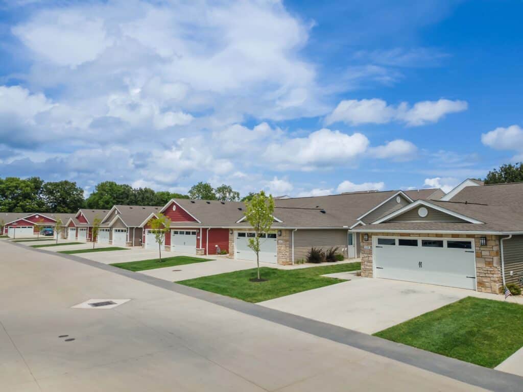 Row of single-story suburban houses with attached garages, neatly trimmed lawns, and clear blue sky overhead.